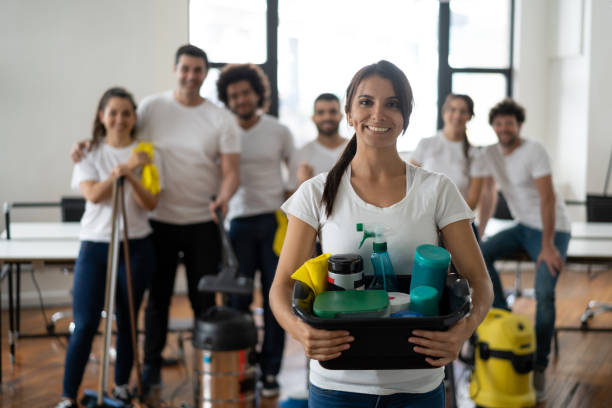 Beautiful cleaning woman with her team at an office holding a bucket with cleaning products all smiling at camera - Service concepts