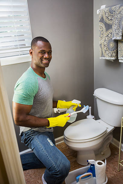 An African American man doing housework.  He is kneeling in the bathroom at the toilet bowl, wearing rubber gloves, with a spray bottle and scrub brush.  He is looking at the camera and smiling.